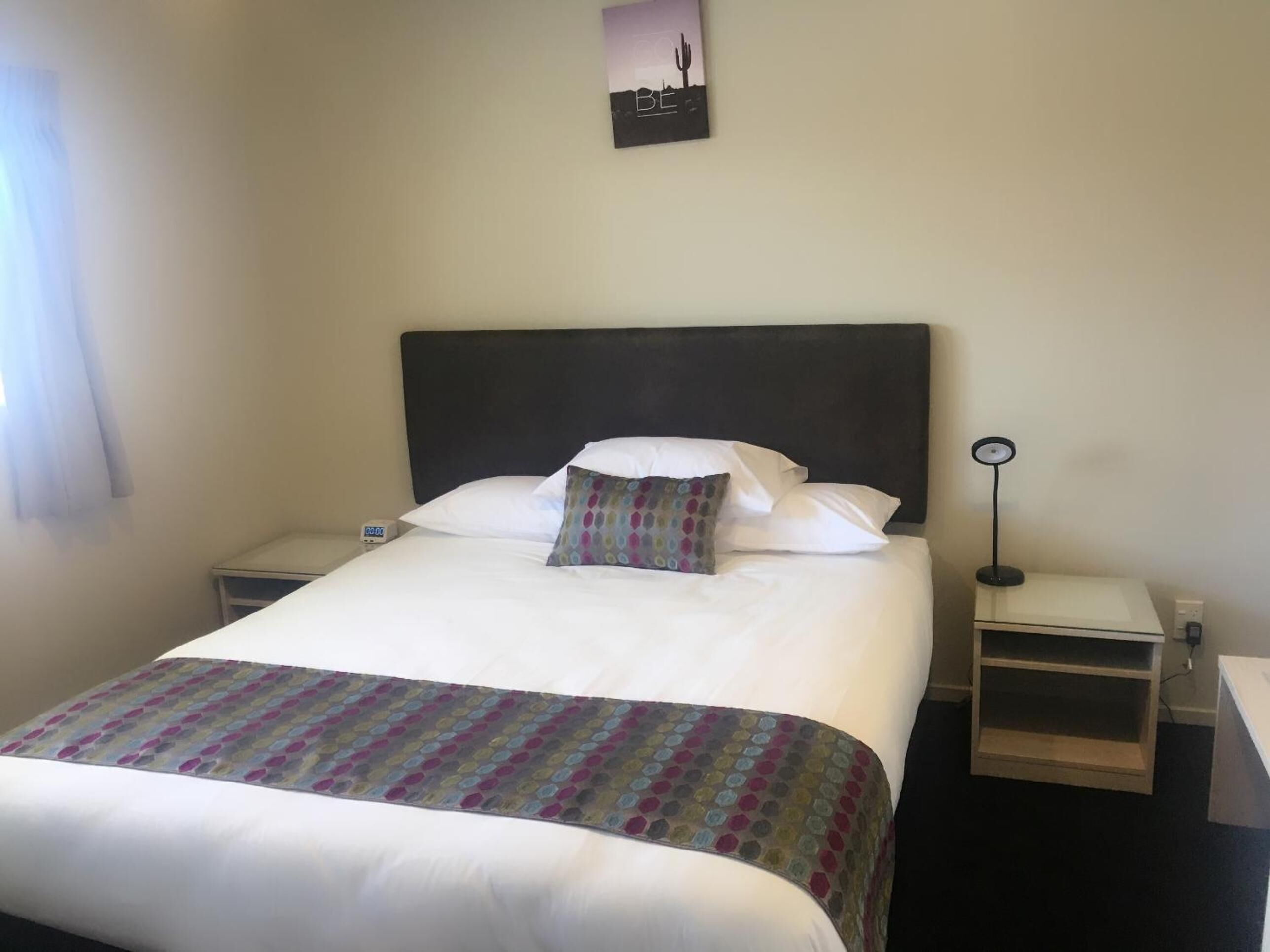 A neatly made bed with a patterned bedspread, white pillows, and a dark headboard. There is a bedside table with a lamp and a small clock. A framed picture hangs on the wall above the bed.