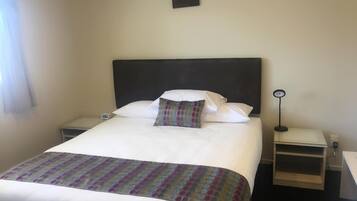 A neatly made bed with a patterned bedspread, white pillows, and a dark headboard. There is a bedside table with a lamp and a small clock. A framed picture hangs on the wall above the bed.