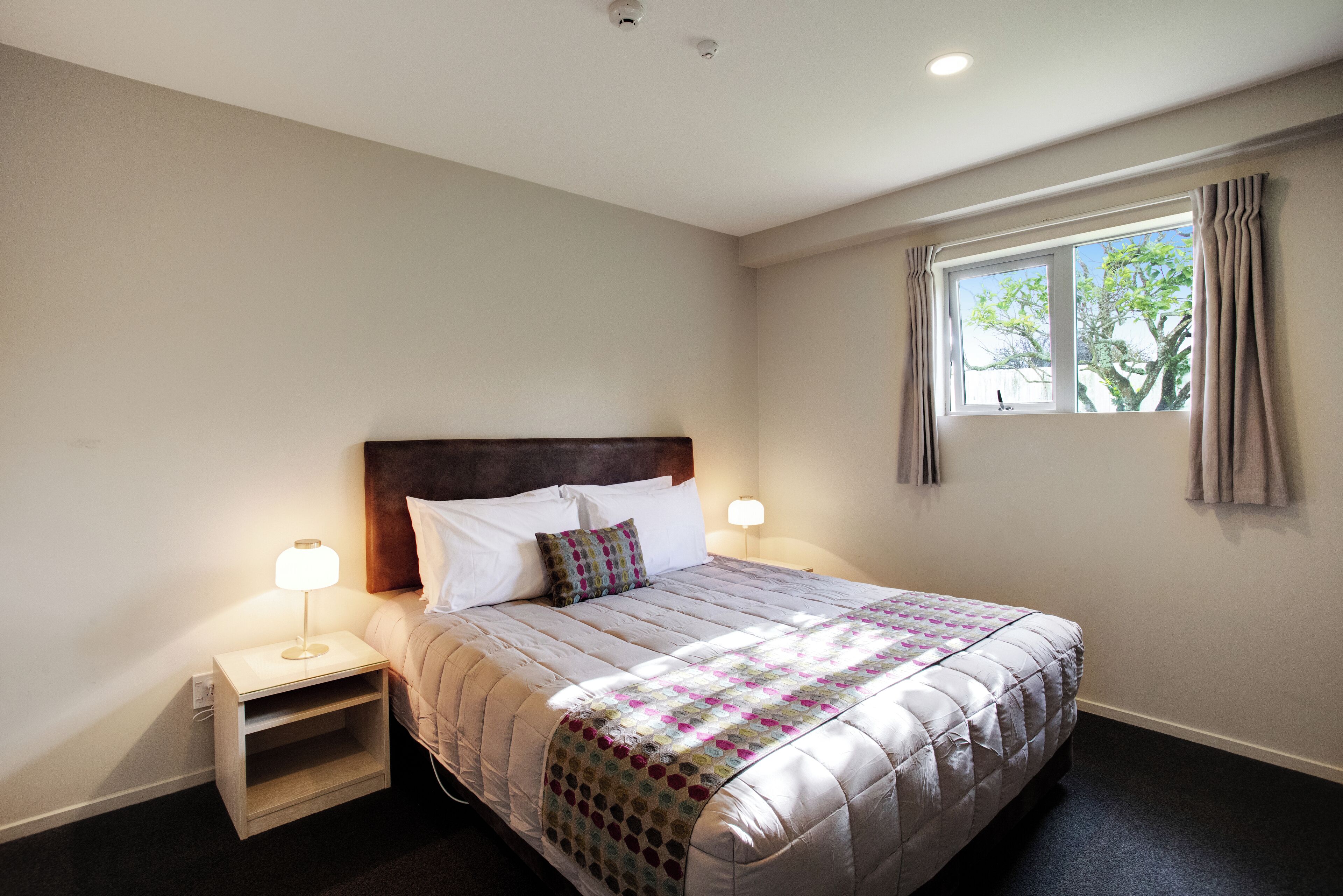 A neatly made bed with a quilt, two bedside lamps, a nightstand, and a window with curtains.