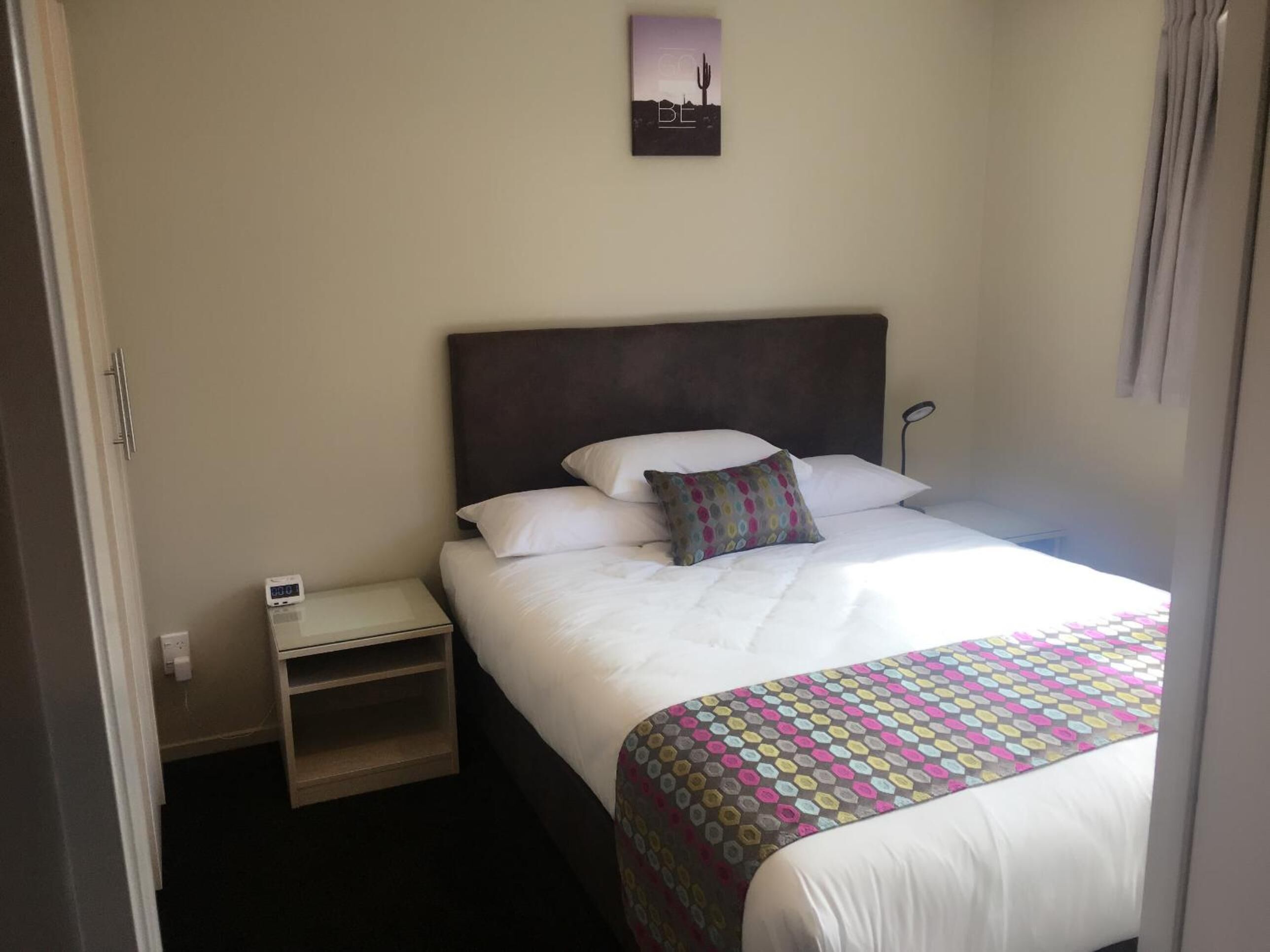 A hotel room with a bed, a bedside table, a nightstand, a wall clock, and a small picture frame.