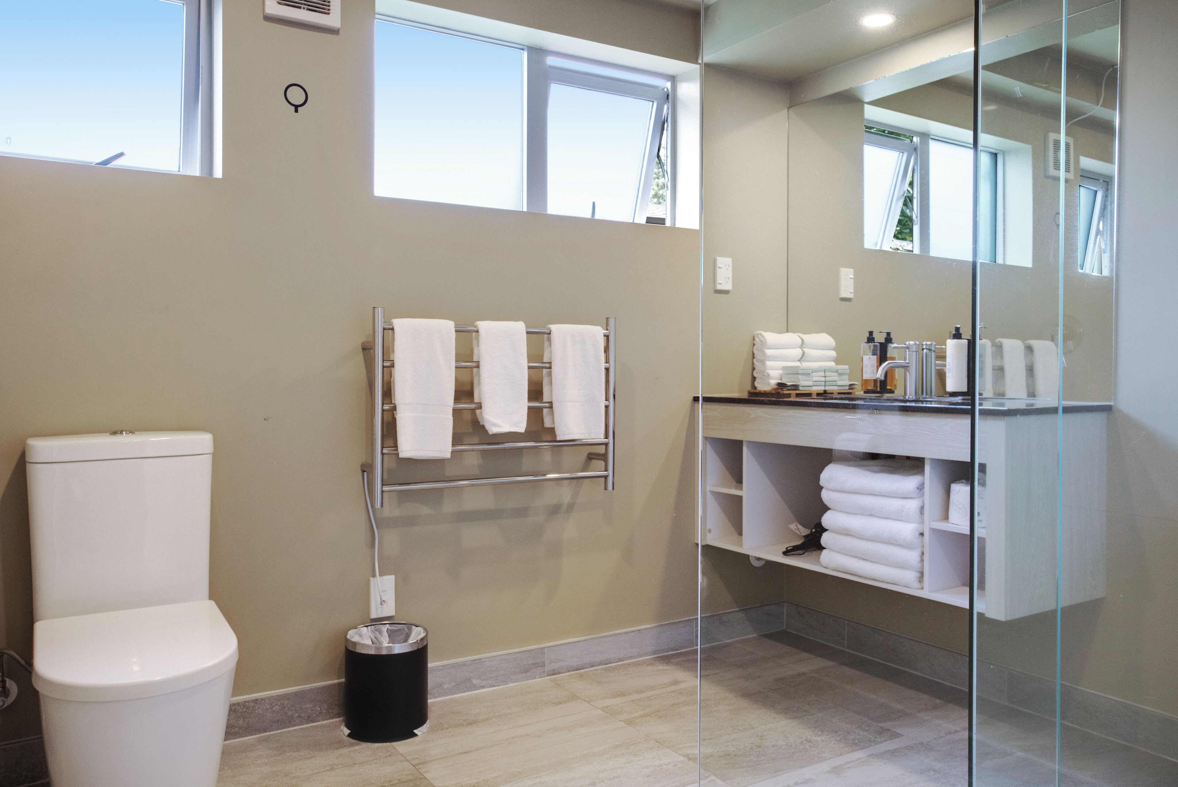 Ein modernes Badezimmer mit Toilette, Handtuchhalter, einer Duschkabine aus Glas und einem Waschtisch mit Pflegeprodukten.