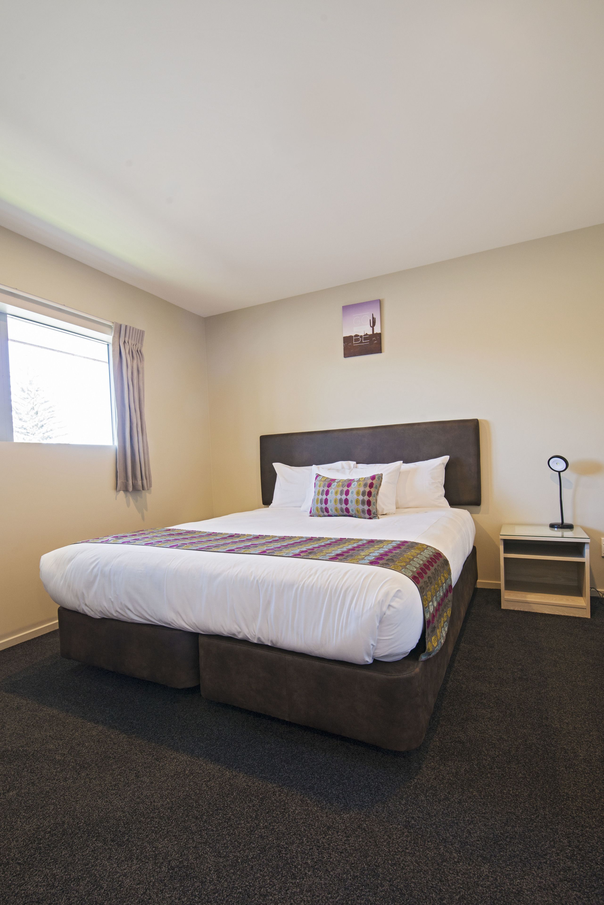 A neatly made bed with a headboard, a bedside table with a lamp, and a window with curtains.