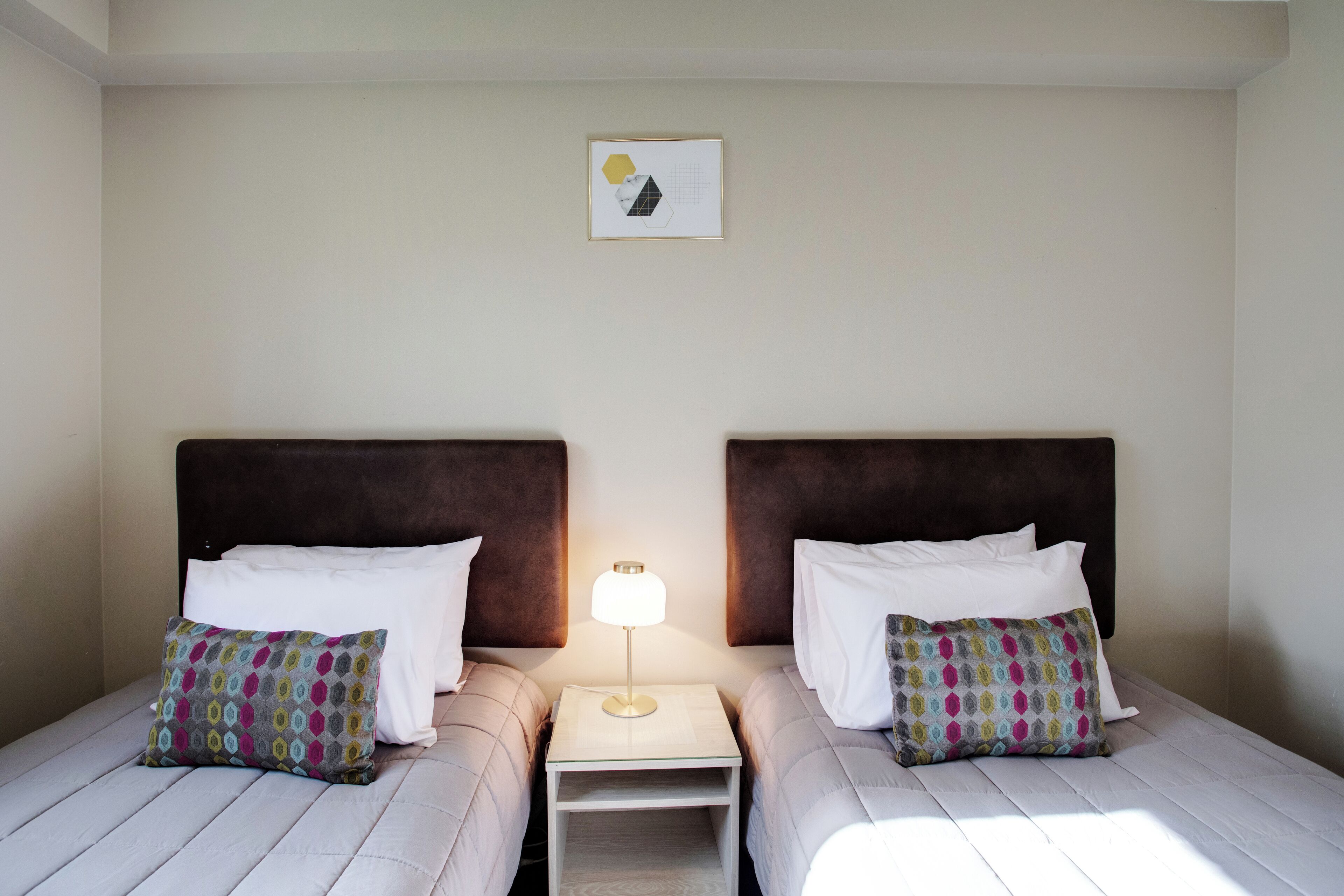 A hotel room with two beds, each with a headboard, a bedside table with a lamp, and a framed picture on the wall.
