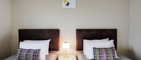 A hotel room with two beds, each with a headboard, a bedside table with a lamp, and a framed picture on the wall.