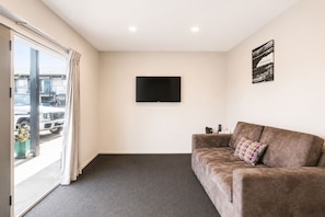 A room with a grey carpet, a brown sofa with a polka-dotted pillow, a flat-screen TV on the wall, and a window with curtains. - Ellerslie Palms Motel (Auckland)
