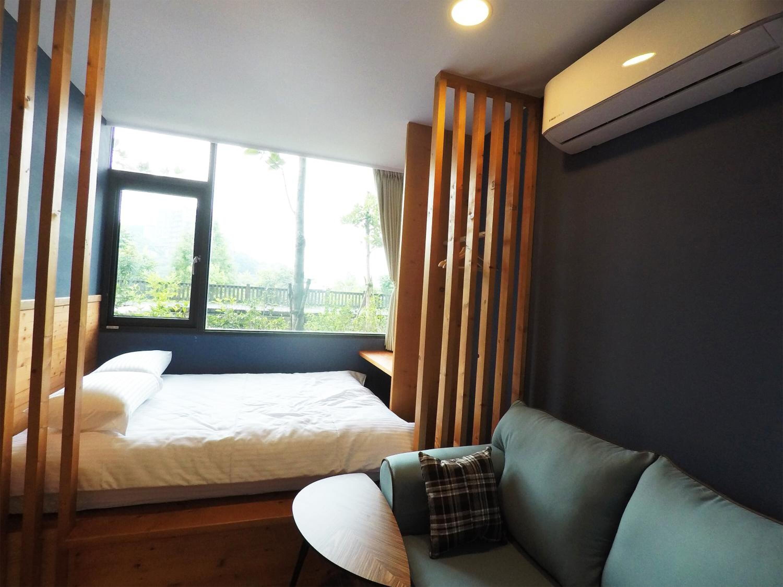 A bedroom with a bed, a green sofa, a window with a view of greenery, and an air conditioning unit.
