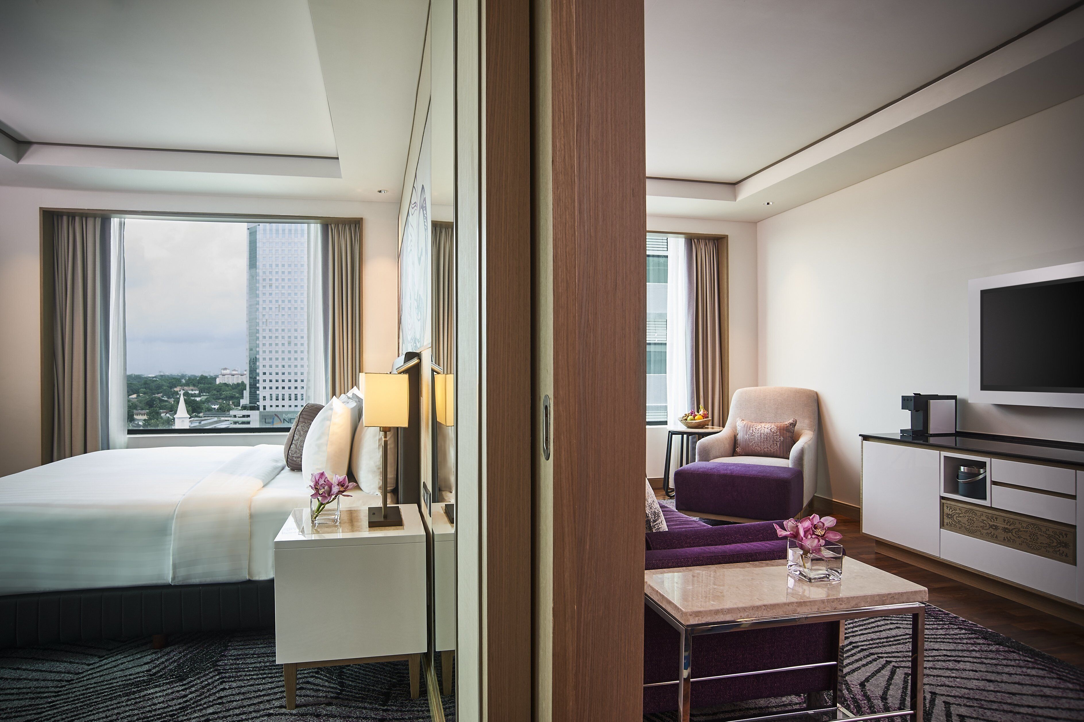 A hotel room with a bed, a desk, a chair, a TV, and a view of a cityscape.
