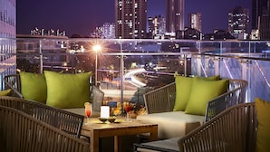 Una terraza en la azotea con mobiliario de mimbre, una mesa con vela y vistas a la ciudad de noche.