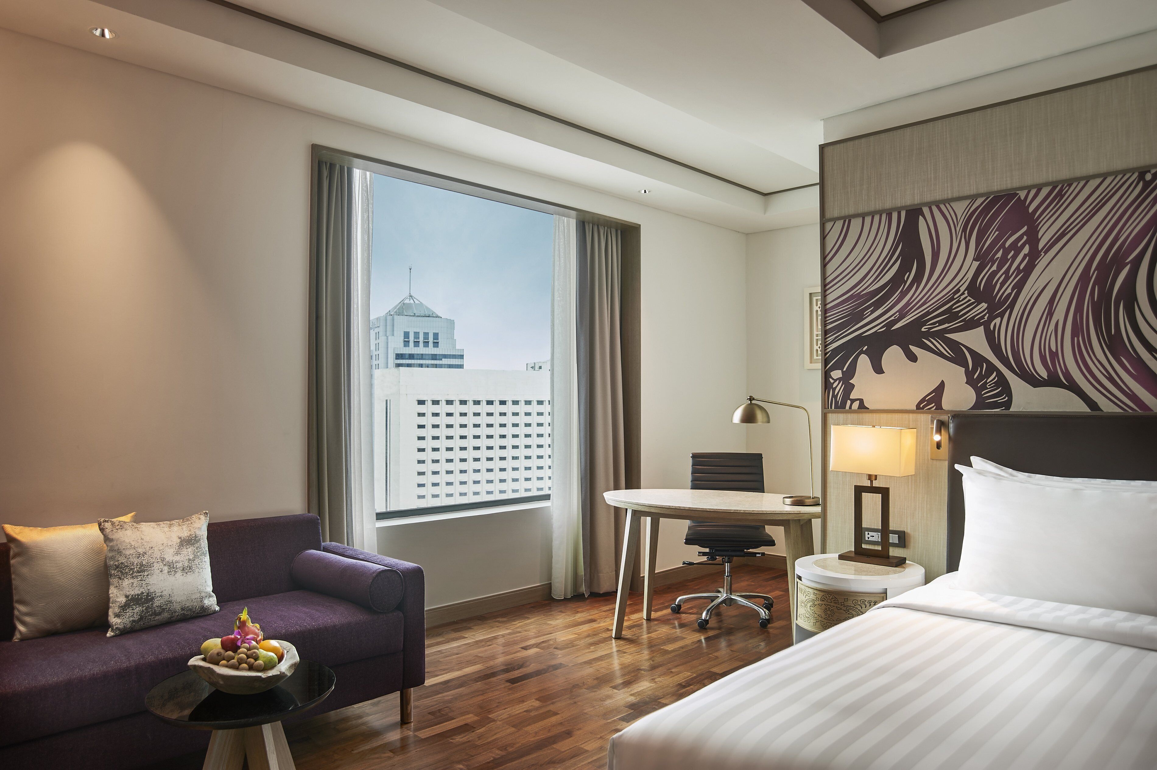 A hotel room with a bed, a sofa, a dining table, a chair, and a small table with fruit.