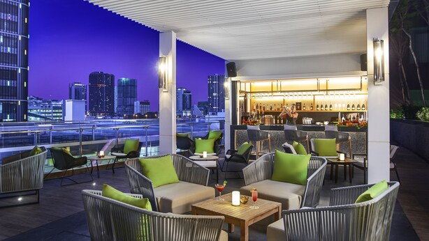 Un bar en la azotea con mesas al aire libre, vistas a la ciudad y una zona de bar con botellas y vasos.
