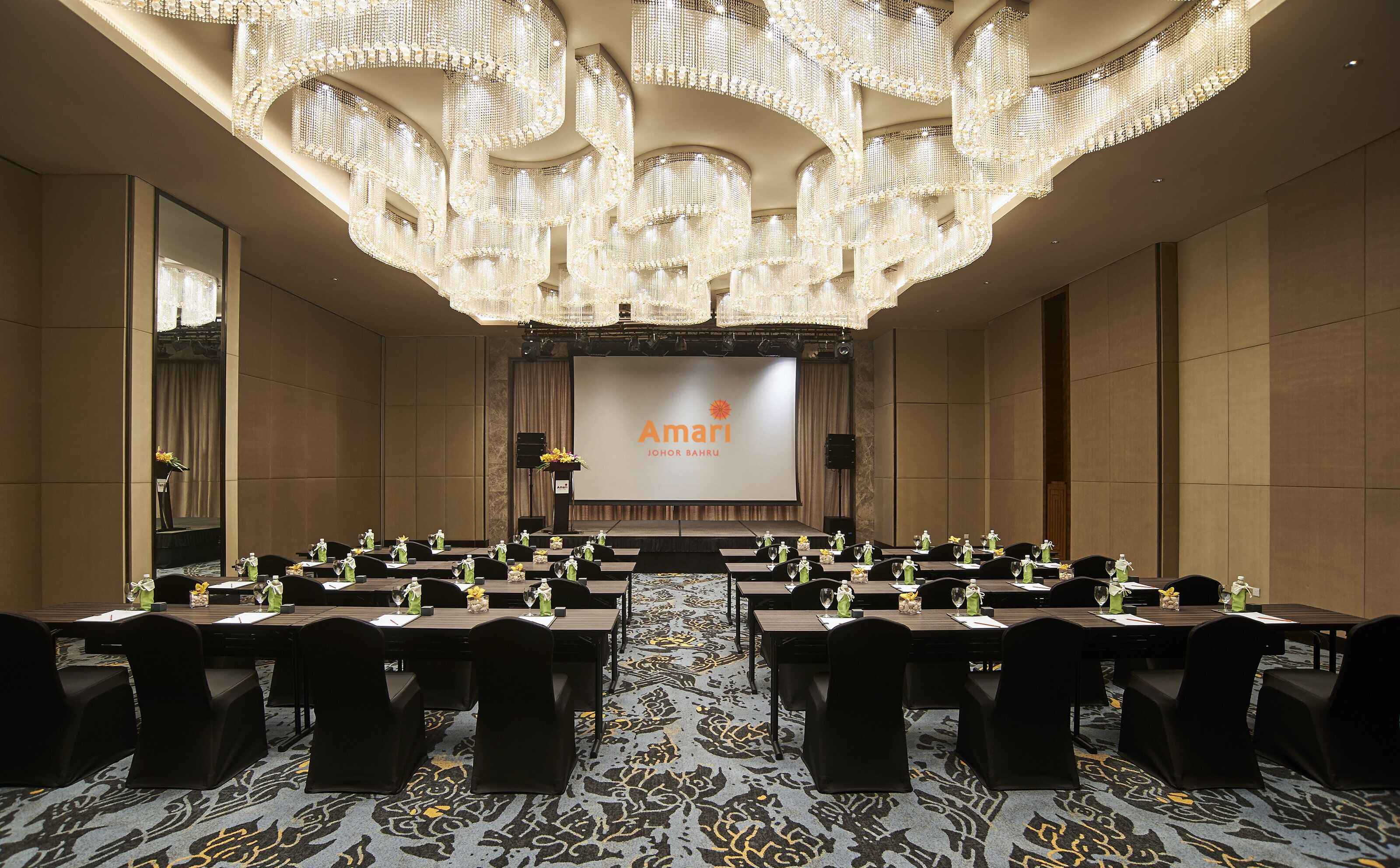 Una sala de conferencias con un escenario, varias mesas y sillas, que incluye un candelabro y una pantalla que muestra el nombre del hotel.