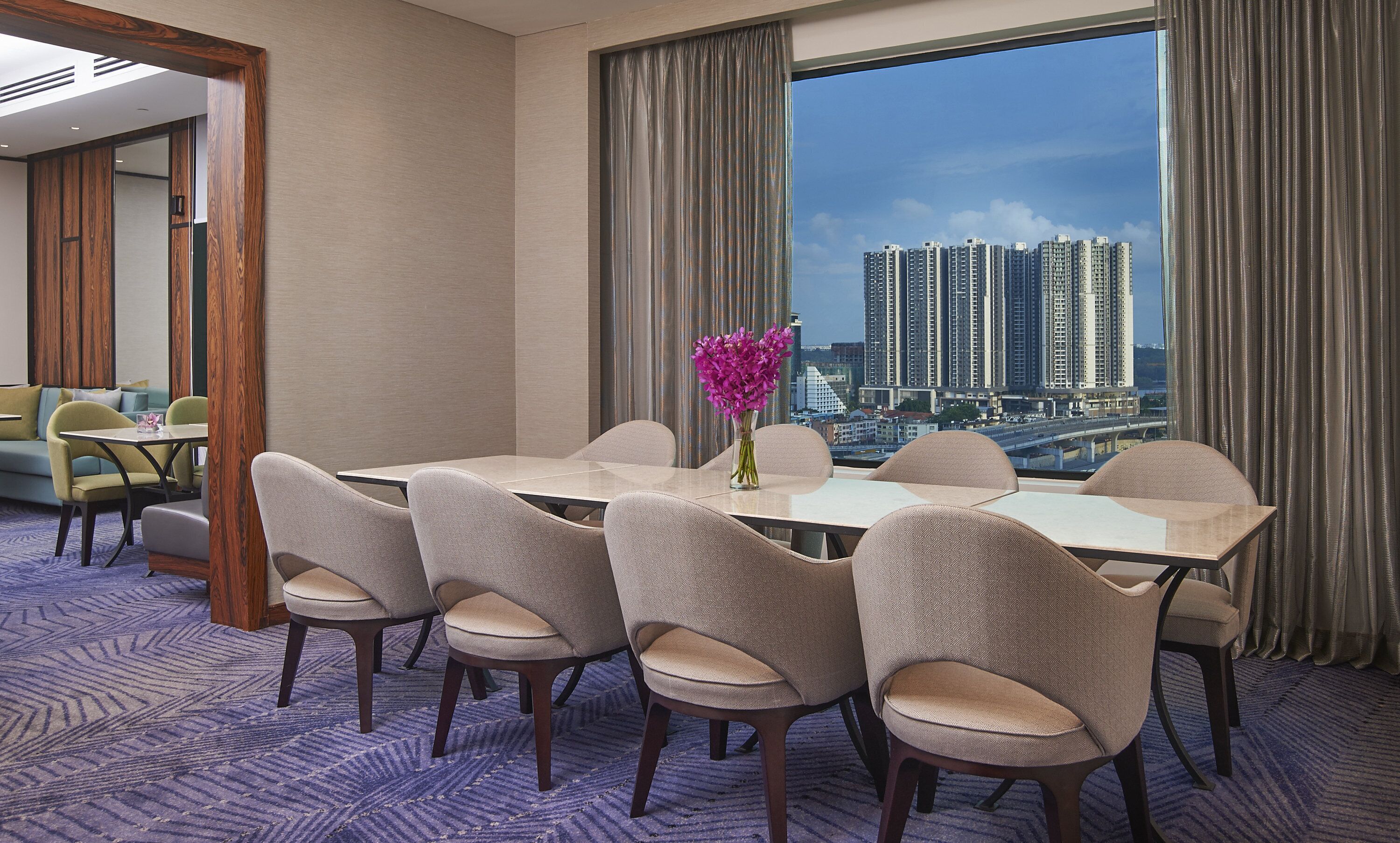 Une chambre d’hôtel dotée d’une grande table à manger, de six chaises, d’un vase rempli de fleurs et offrant une vue sur la ville grâce à de grandes fenêtres.