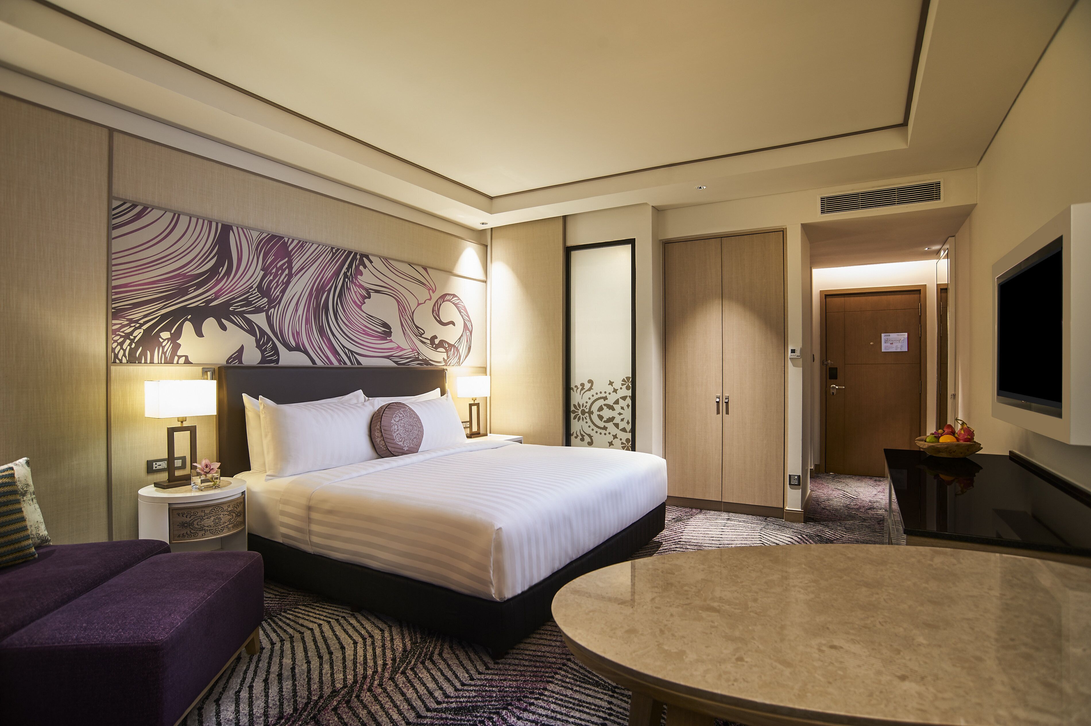 A hotel room with a large bed, a small round table, a sofa, a TV, and a decorative wall piece.