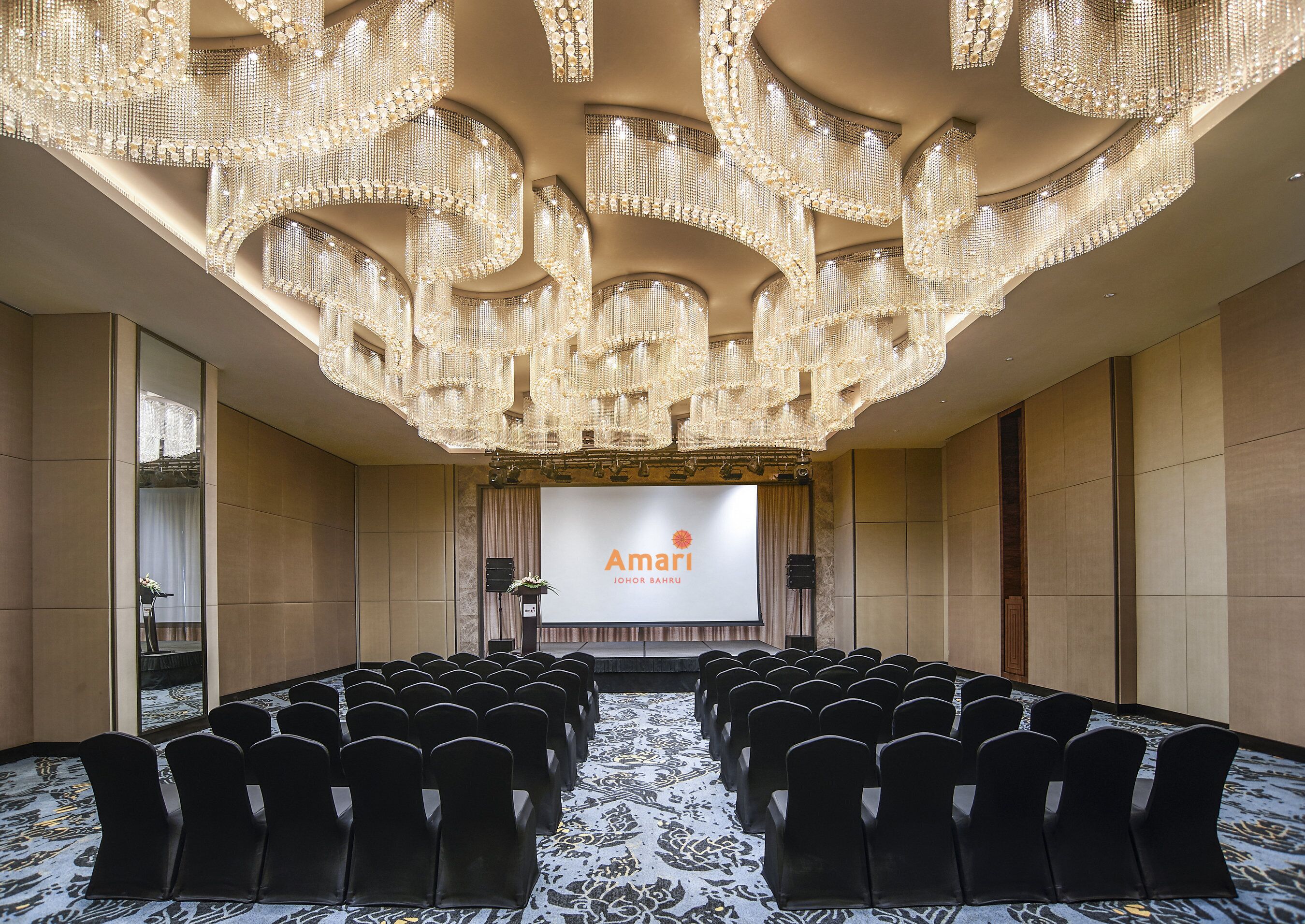 A conference room with rows of black chairs, a large screen displaying 'Amari JOHOR BAHRU', and ornate chandeliers.