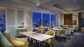 A modern hotel lobby with a city view, featuring a dining area with tables and chairs, a sofa, and a small table with a vase.