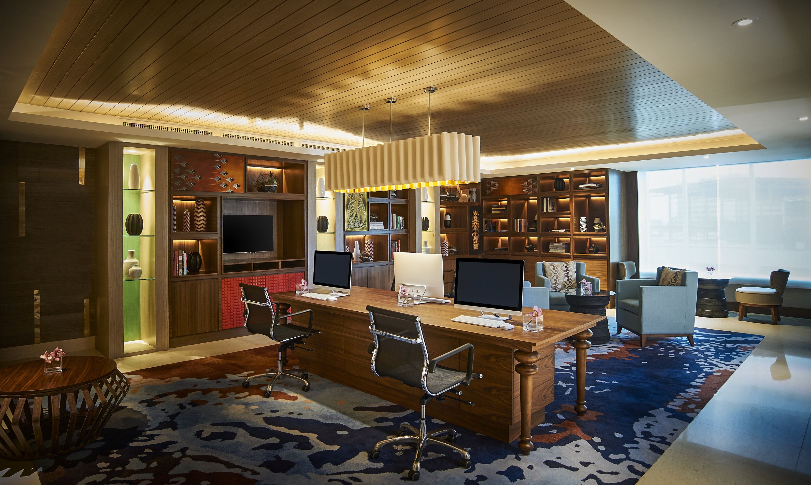 A modern office space with a wooden desk, two computer monitors, a television, and a large bookshelf.