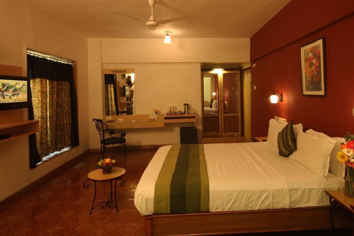 Une chambre d’hôtel comprenant un lit, une table de chevet, une lampe, un miroir et une petite table sur laquelle est posé un vase de fleurs.