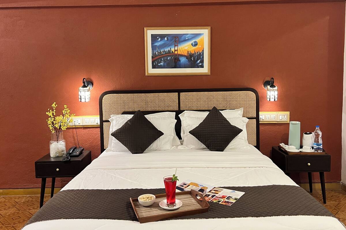 Une chambre d’hôtel avec un lit, une table de chevet avec un téléphone, un plateau avec des en-cas, un verre d’une boisson rouge et un tableau encadré au mur.