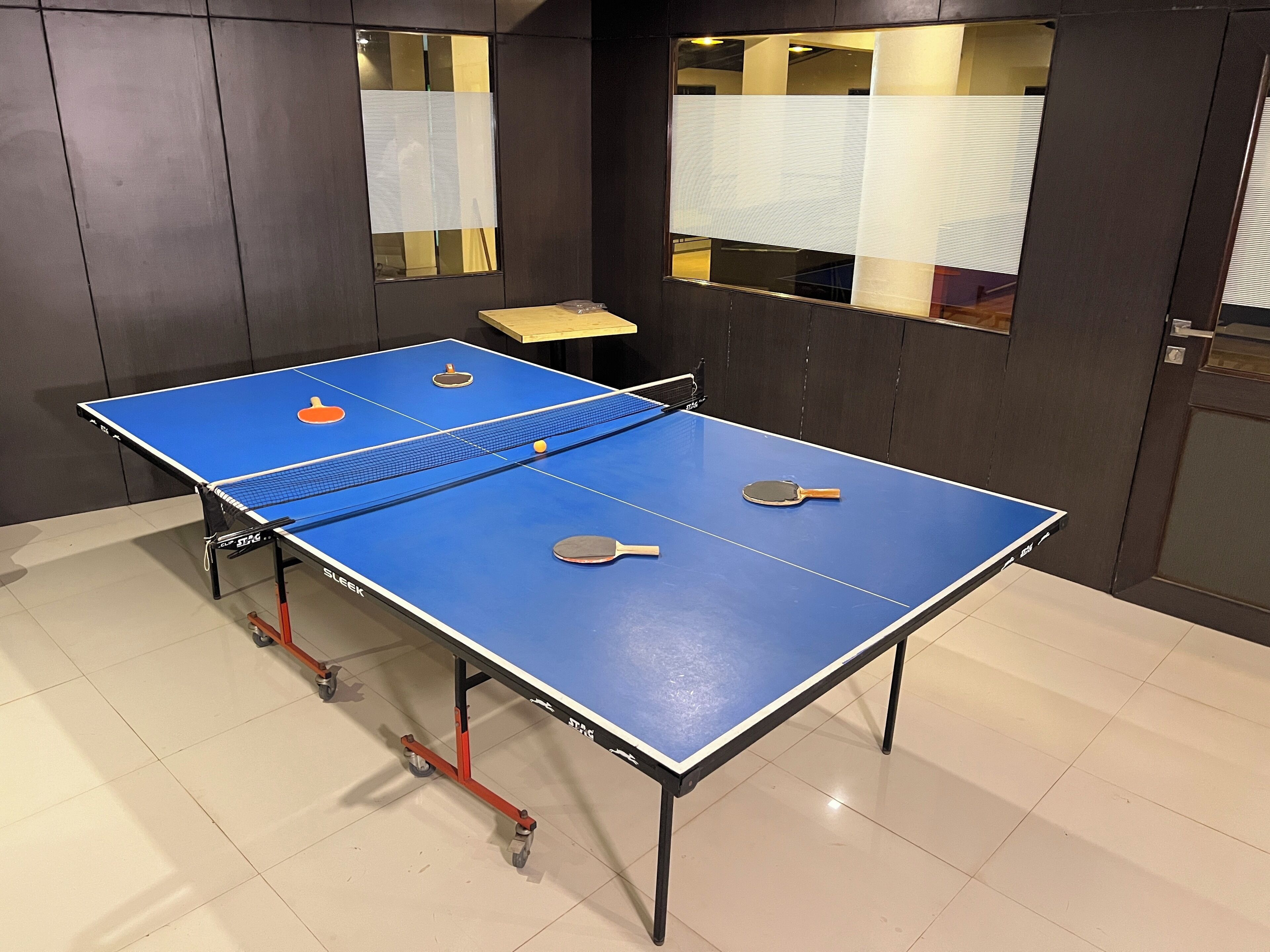 Una mesa de ping pong con dos paletas y una pelota, frente a una pared oscura con ventanas.