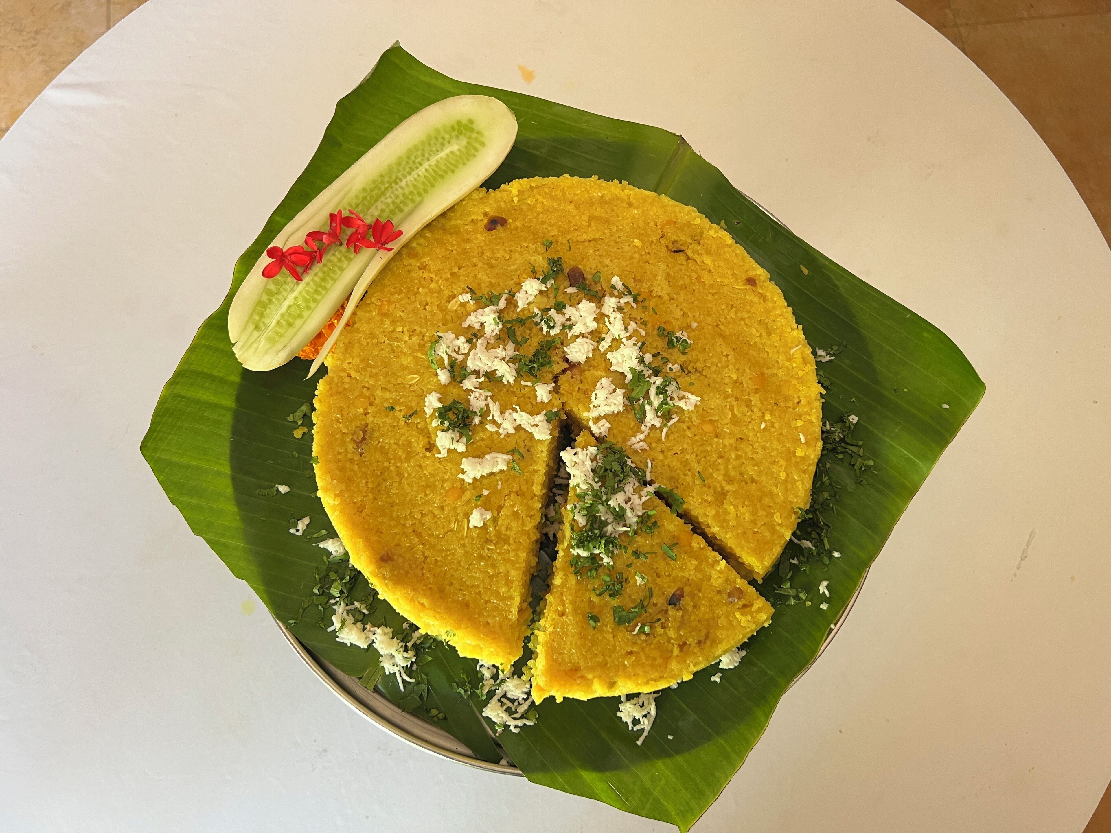 Un plat de riz jaune garni d’herbes aromatiques et de noix de coco râpée, servi sur une feuille de bananier, accompagné de concombre et de fleurs rouges.