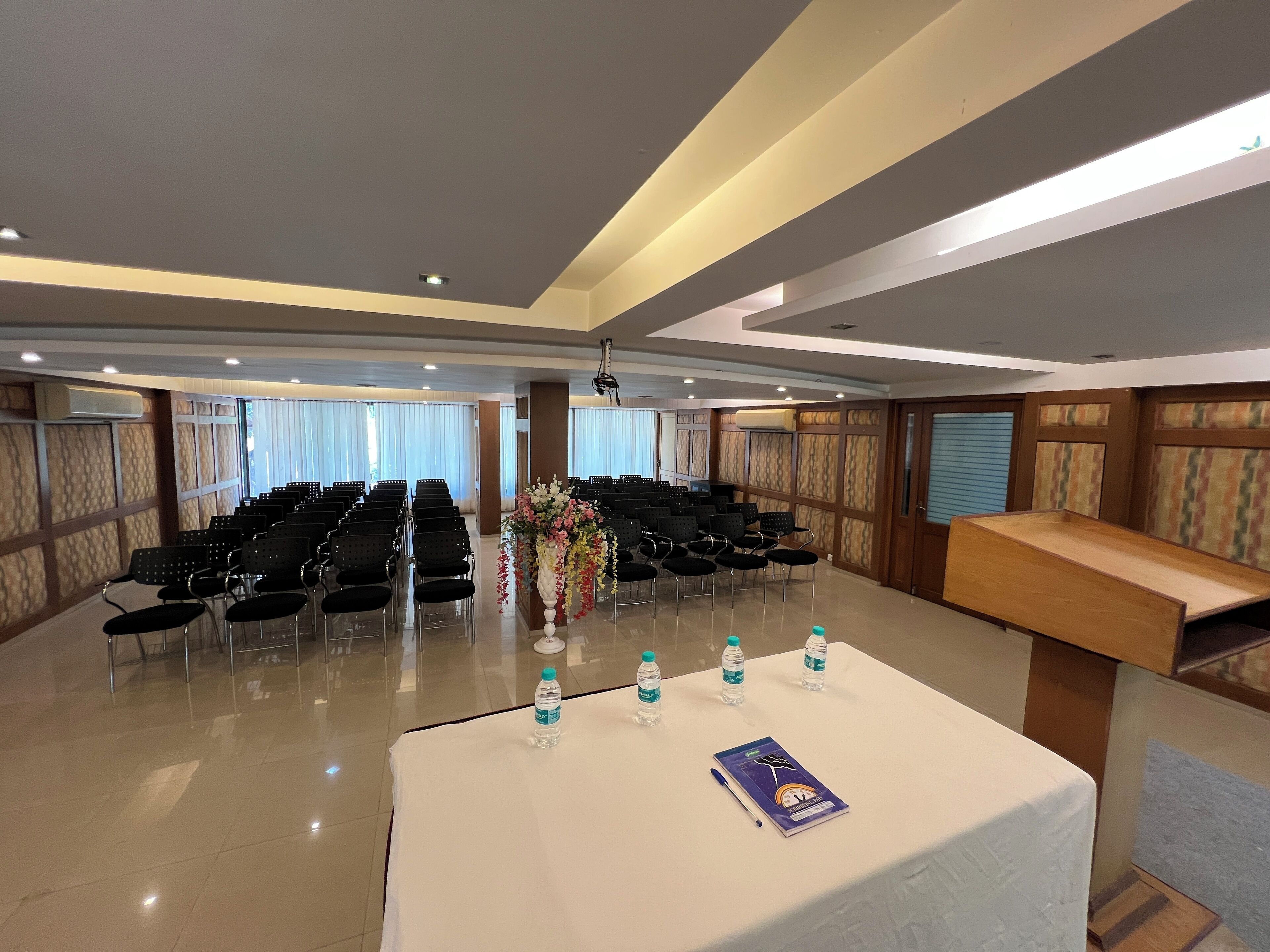 Una sala conferenze con file di sedie, un tavolo con bottiglie d'acqua e un quaderno, e un composizione floreale.