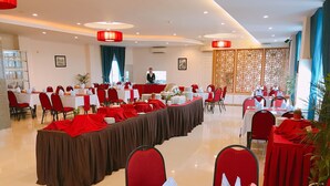 Um salão de banquetes com várias mesas redondas, cadeiras vermelhas e uma mesa longa com toalha marrom.