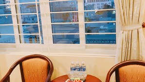 Quarto com duas cadeiras de madeira, uma pequena mesa redonda com água engarrafada e petiscos, e uma janela com vista para edifícios.