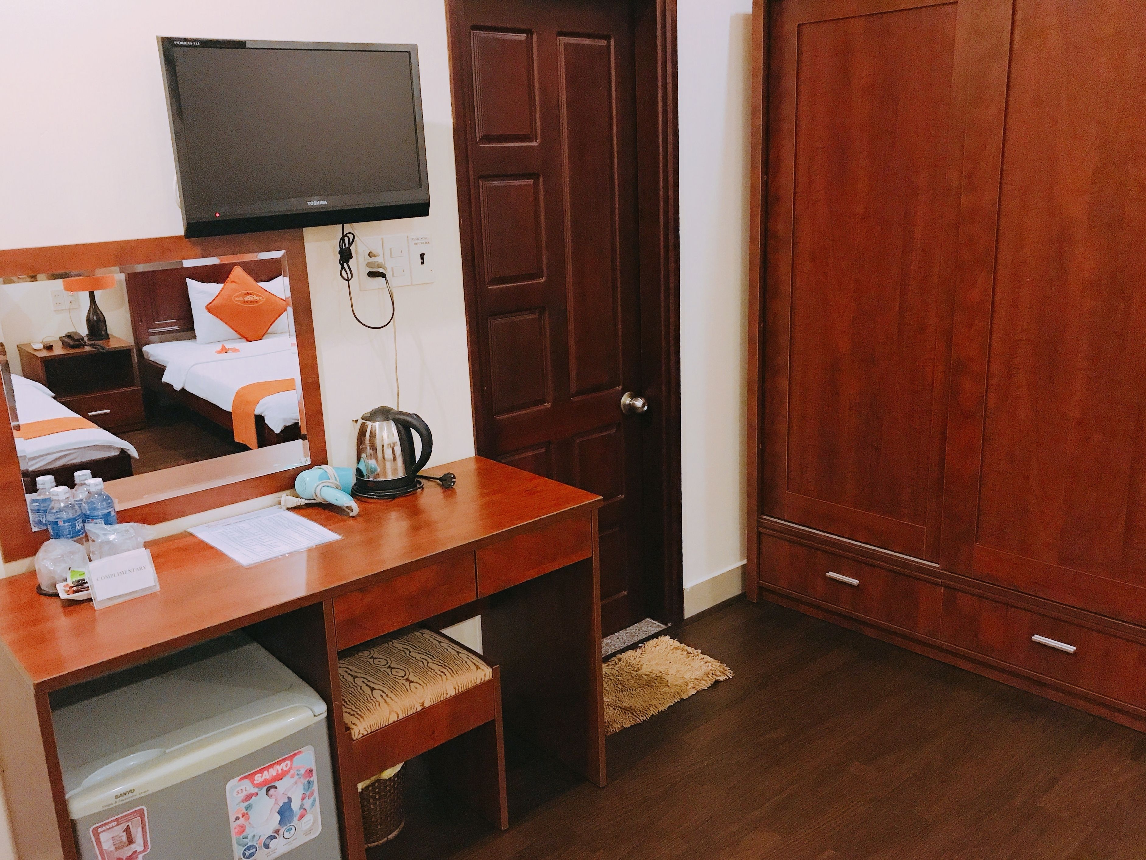 Une chambre d’hôtel avec un bureau en bois, un téléviseur fixé au-dessus, un lit avec des oreillers orange, une bouilloire et un mini-bar.
