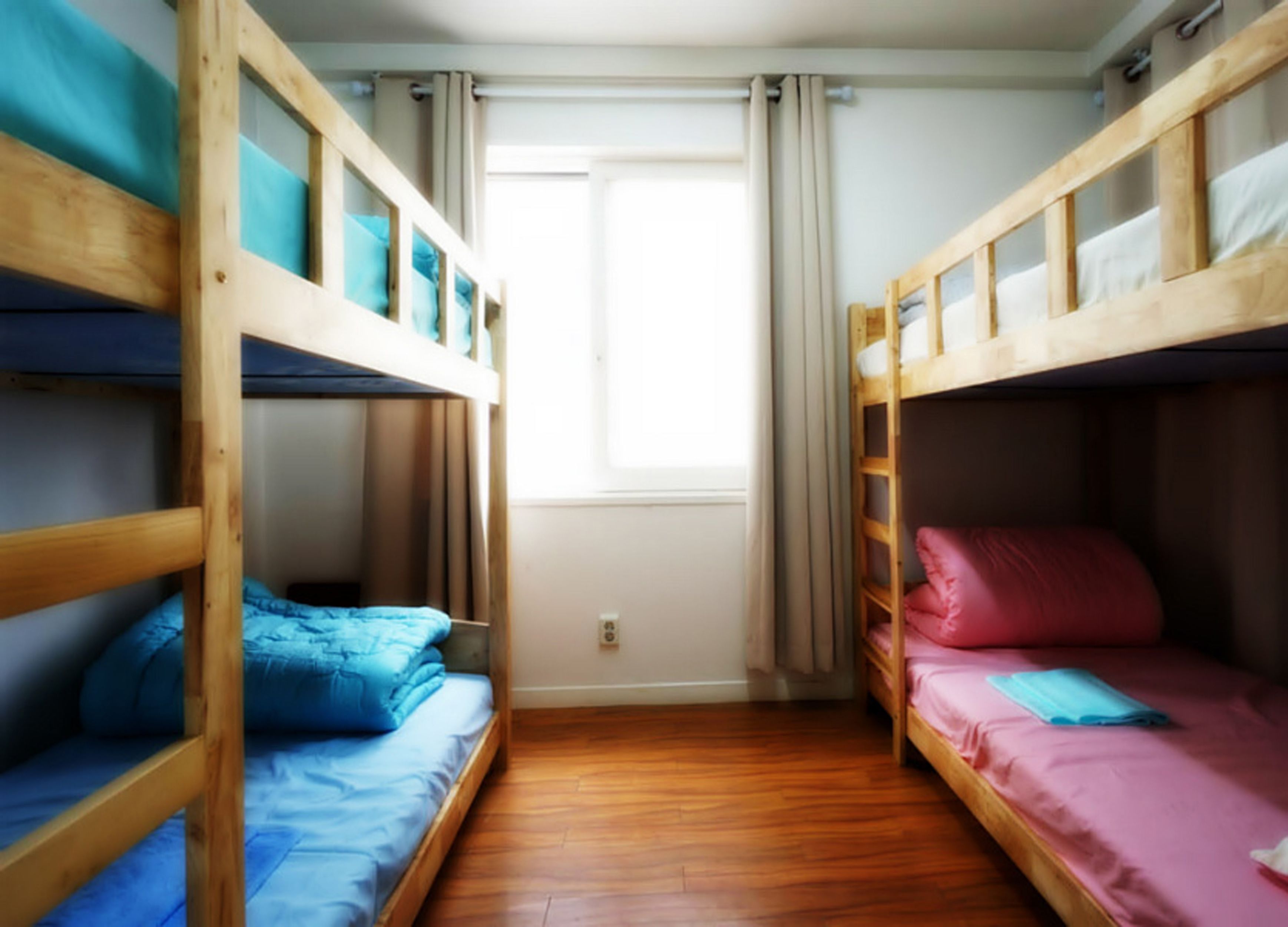 A room with two bunk beds, wooden flooring, a window with curtains, and a plain wall.