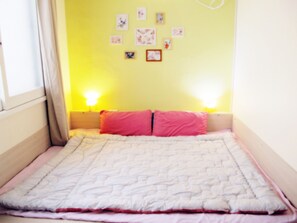 Una habitación pequeña con cama individual, pared amarilla, colcha blanca y almohadas rosas. Cuenta con luces fijadas a la pared y varias fotografías pequeñas enmarcadas.