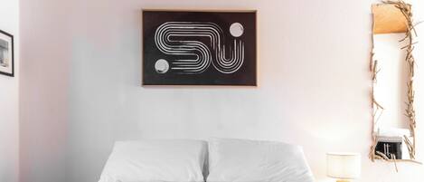 Un dormitorio con una cama, una obra de arte abstracta colgada en la pared, una lámpara de noche y un espejo también sujeto a la pared.