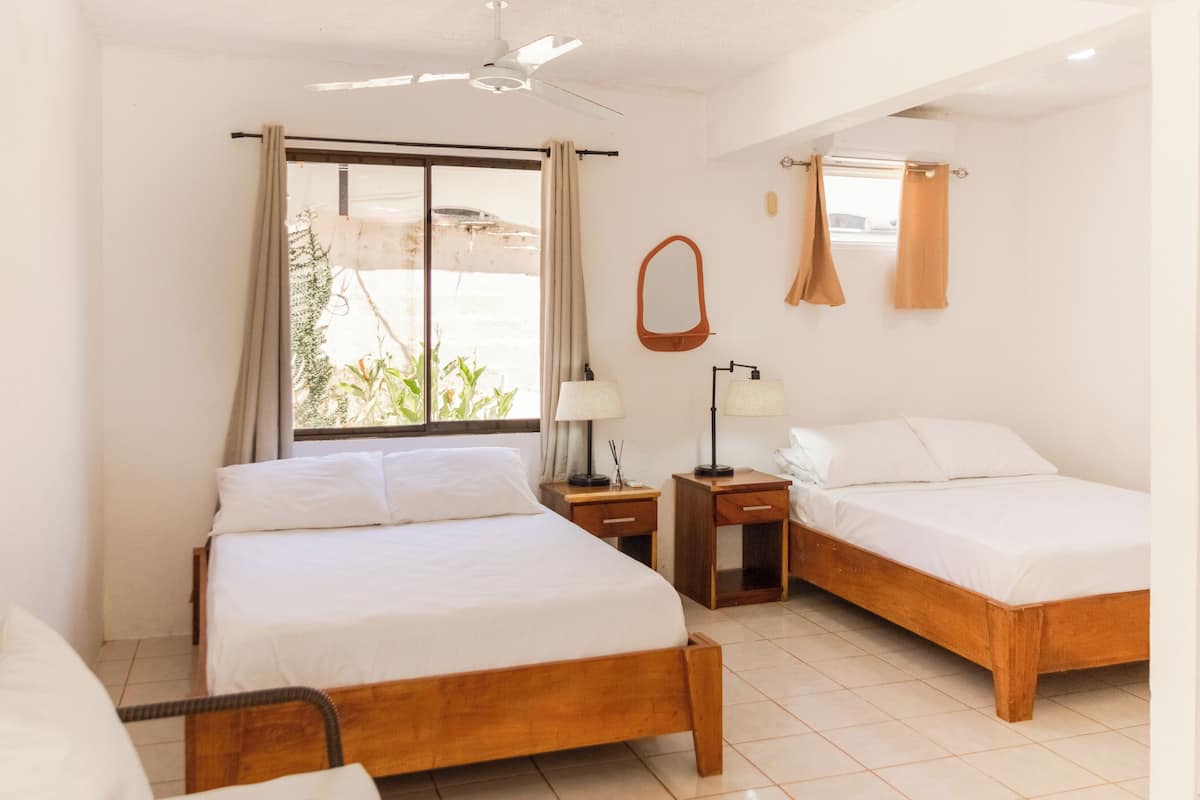 Una habitación de hotel con dos camas de madera, un ventilador de techo, una ventana con cortinas, una mesita de noche con lámpara y un espejo en la pared.