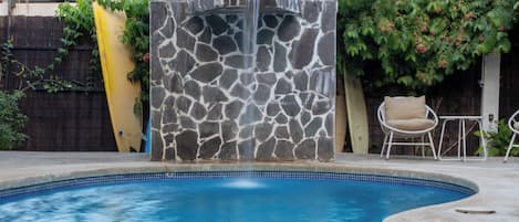 Une piscine agrémentée d’une cascade en pierre, entourée d’une clôture en bois et de plantes.