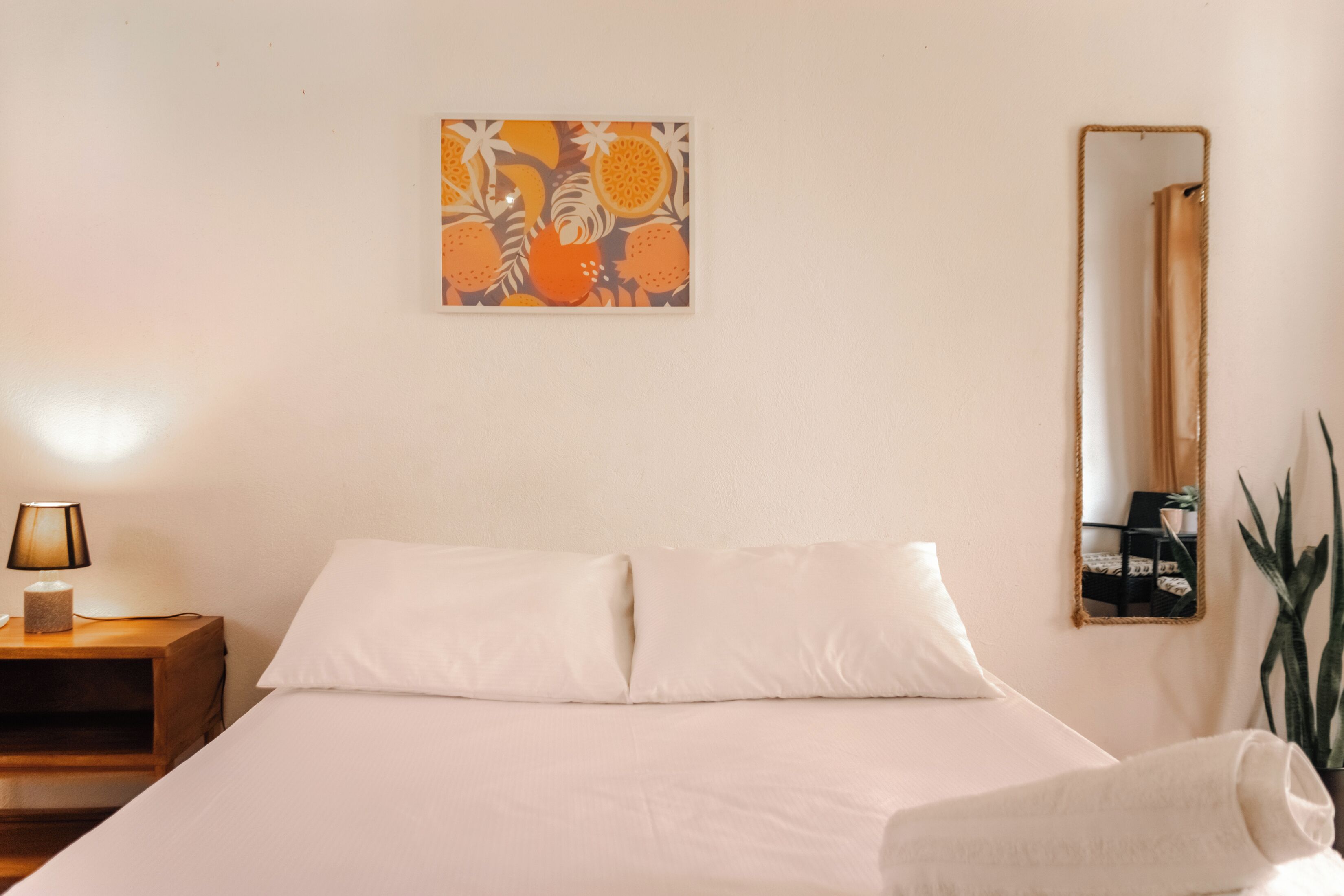 Cama bem arrumada com lençóis brancos, abajur, espelho fixado na parede e uma obra de arte emoldurada sobre a cama.