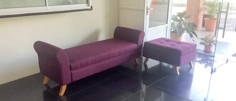Un área de recepción con un sofá y reposapiés morados, una puerta de cristal y plantas en macetas.
