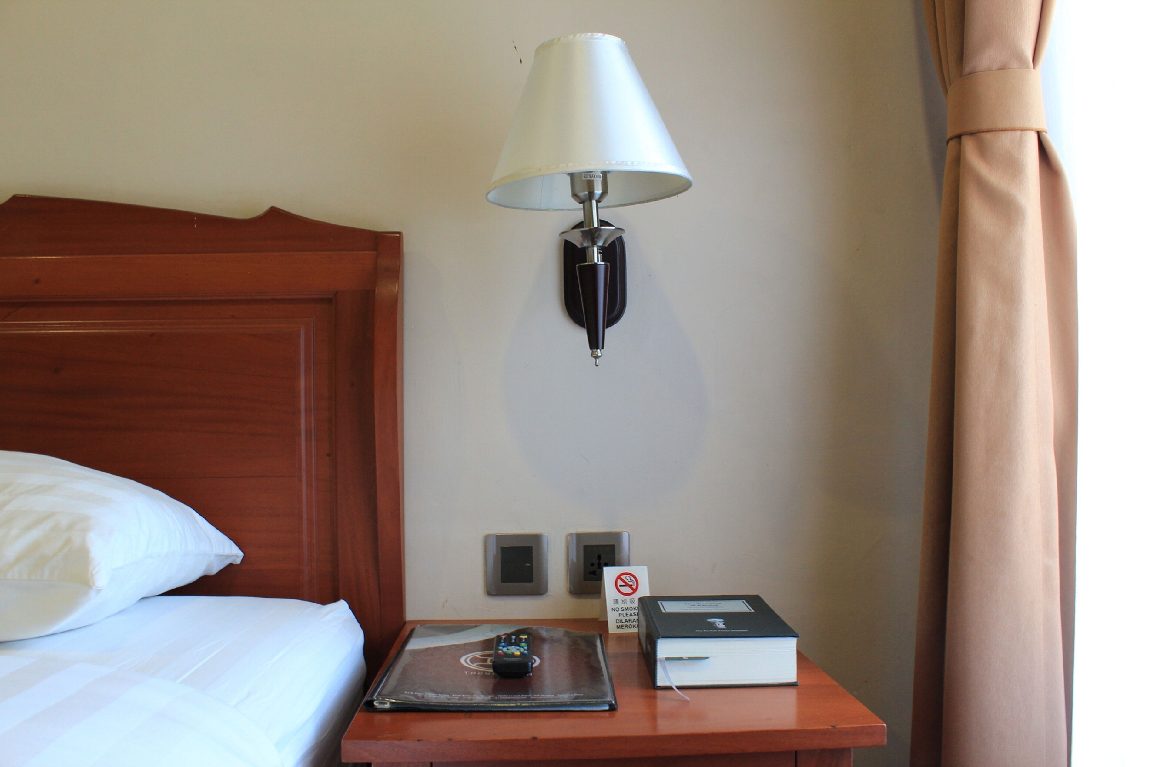 Habitación de hotel con cabecera de madera, mesita de noche con lámpara, cartel que prohíbe fumar y un libro sobre la mesa.