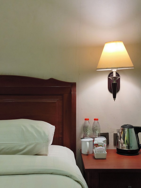 A hotel room with a bed, a nightstand with a lamp, two bottles, a coffee maker, and a kettle.