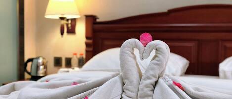 Una cama bien hecha con toallas dispuestas en forma de corazón, una lámpara de noche y una tetera sobre la mesita.
