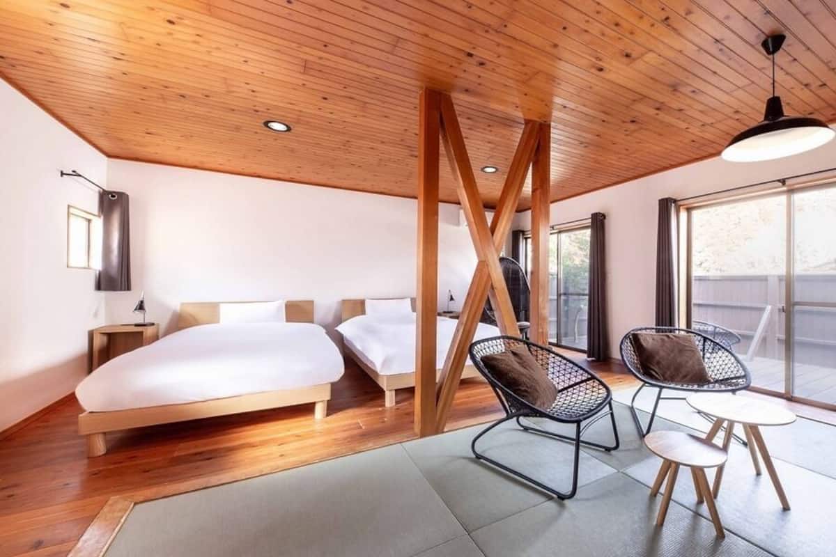 Una habitación de hotel moderna con techo de madera, una cama grande, una zona de estar con sillones y un ventanal amplio.
