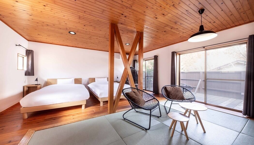 Quarto de hotel moderno com teto de madeira, uma cama, área de estar com cadeiras e uma janela grande.