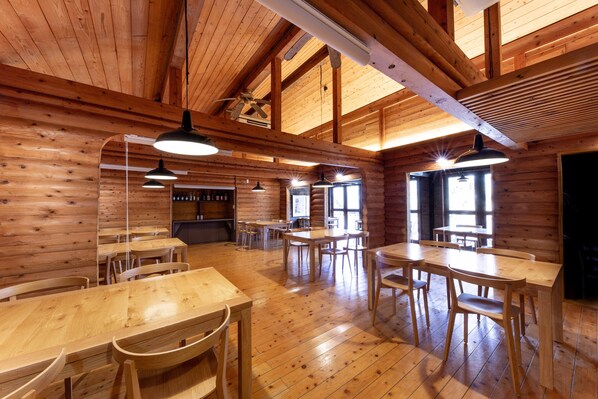 A wooden interior with multiple tables and chairs, a ceiling fan, and large windows allowing natural light. - Ryokan Hanagokoro (Minamioguni)