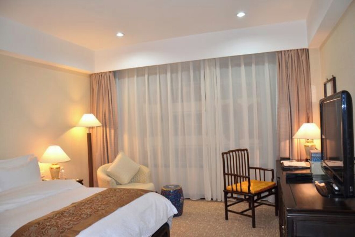 A hotel room with a bed, two chairs, a desk with a computer, a television, and a large window with curtains.