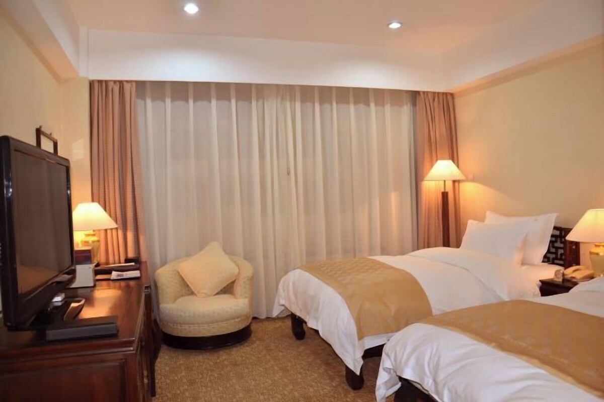 A hotel room with two beds, a television, a chair, and lamps.