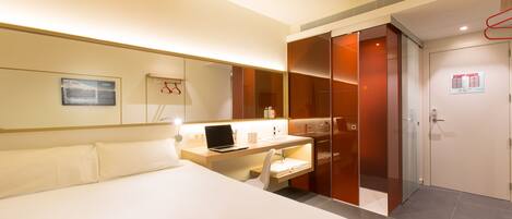 A modern hotel room with a bed, a desk with a laptop, a chair, a wardrobe, and a bathroom.
