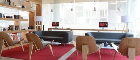 A modern living room with a grey sofa, wooden chairs, and a red rug. There are two screens on the wall and a bookshelf with various items.