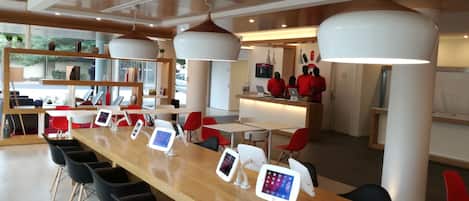 Un área de recepción moderna con un mostrador de madera largo, múltiples sillas rojas y un mostrador con personal uniformado de rojo.