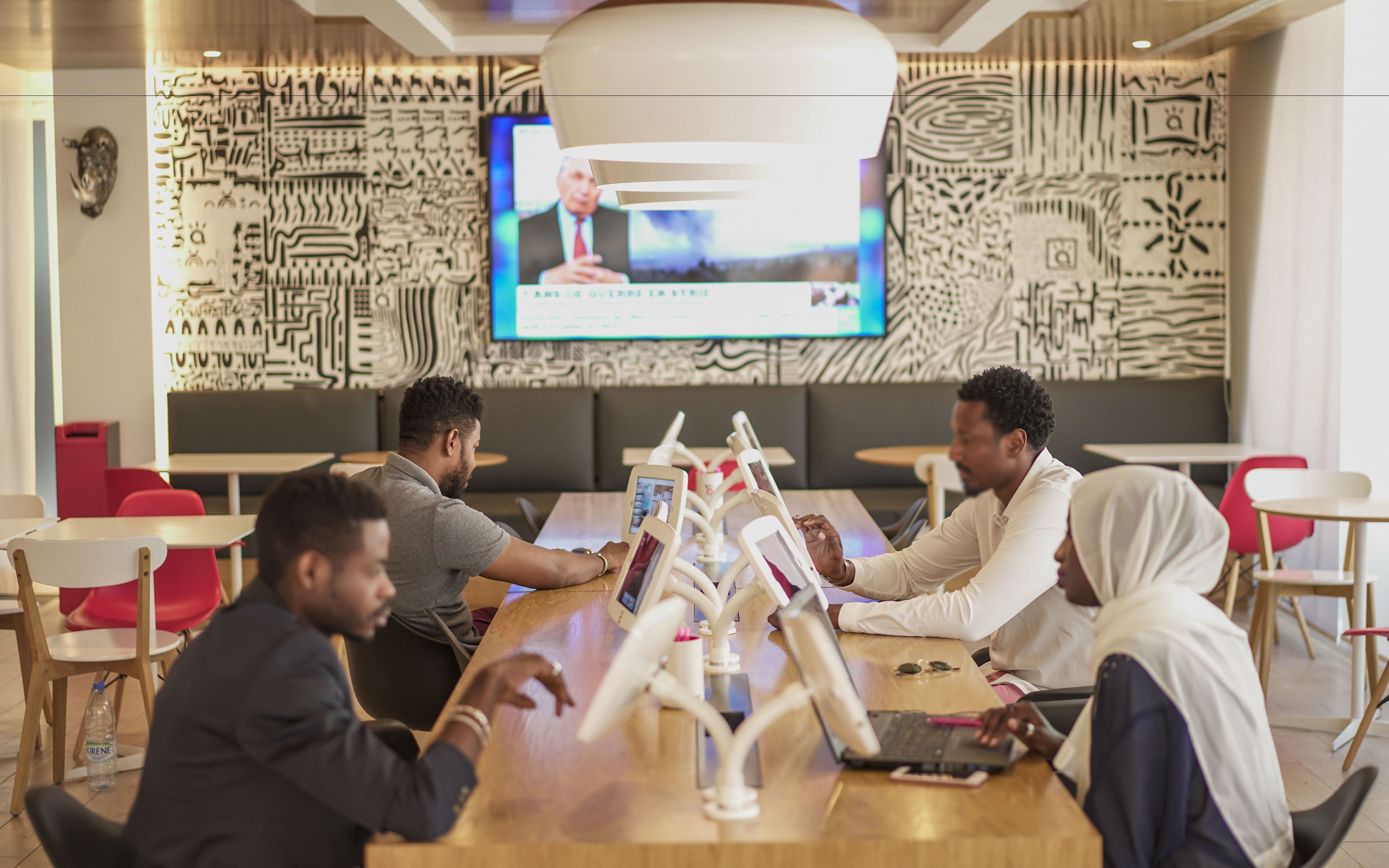 A modern meeting room with a large screen displaying a video, wooden tables, and red chairs.