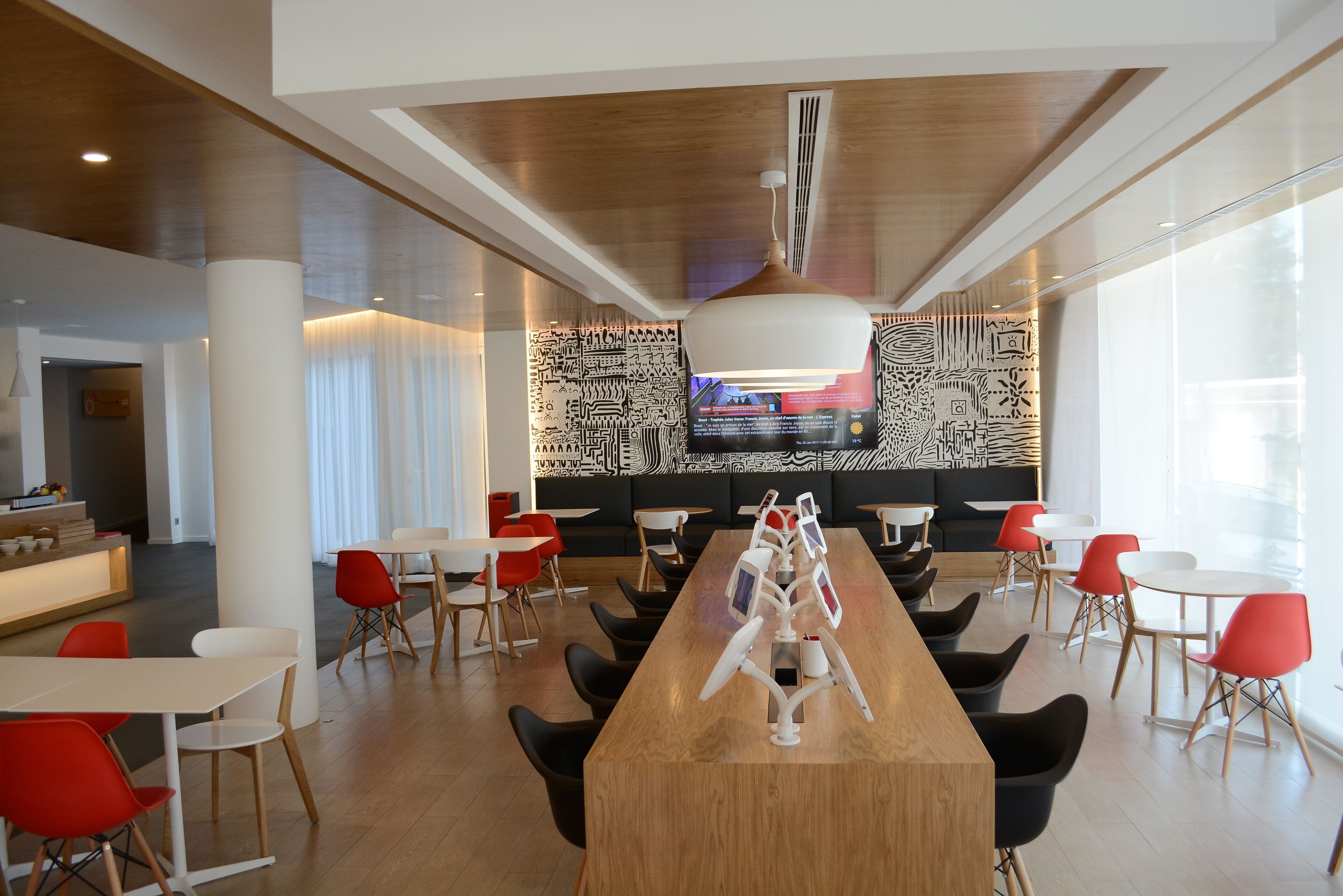 A modern restaurant with a long wooden table, red and black chairs, and a patterned wall.
