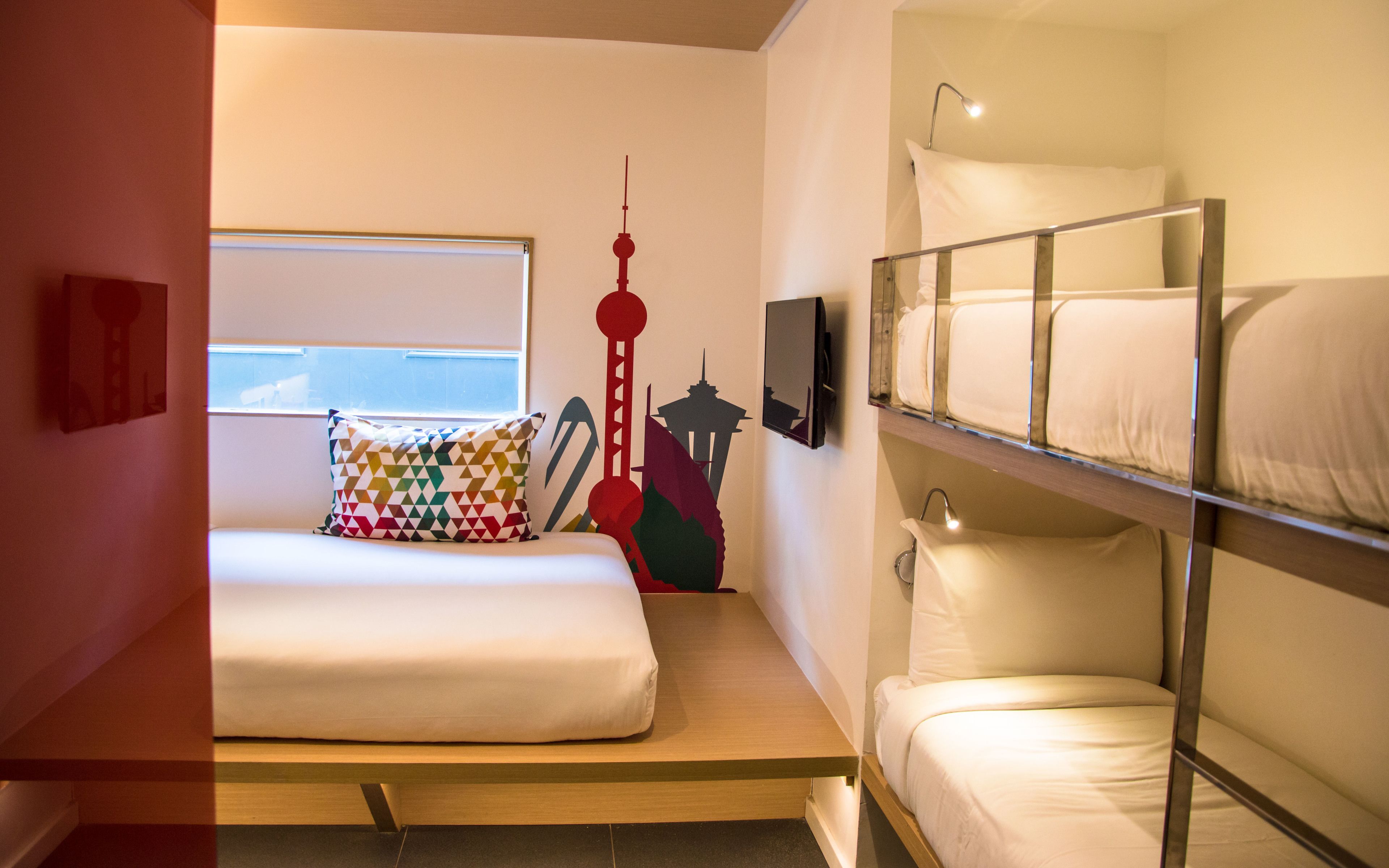 A compact hotel room with a bunk bed, a single bed, a colorful pillow, a wall-mounted TV, and a window with blinds.