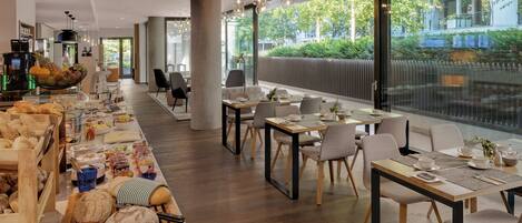 A modern dining area with wooden tables and chairs, a buffet with various breads and pastries, and large windows offering a view of greenery and buildings.